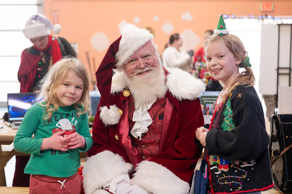 Santa Claus with two smiling kids in holiday sweaters at the Trackers Holiday Faire