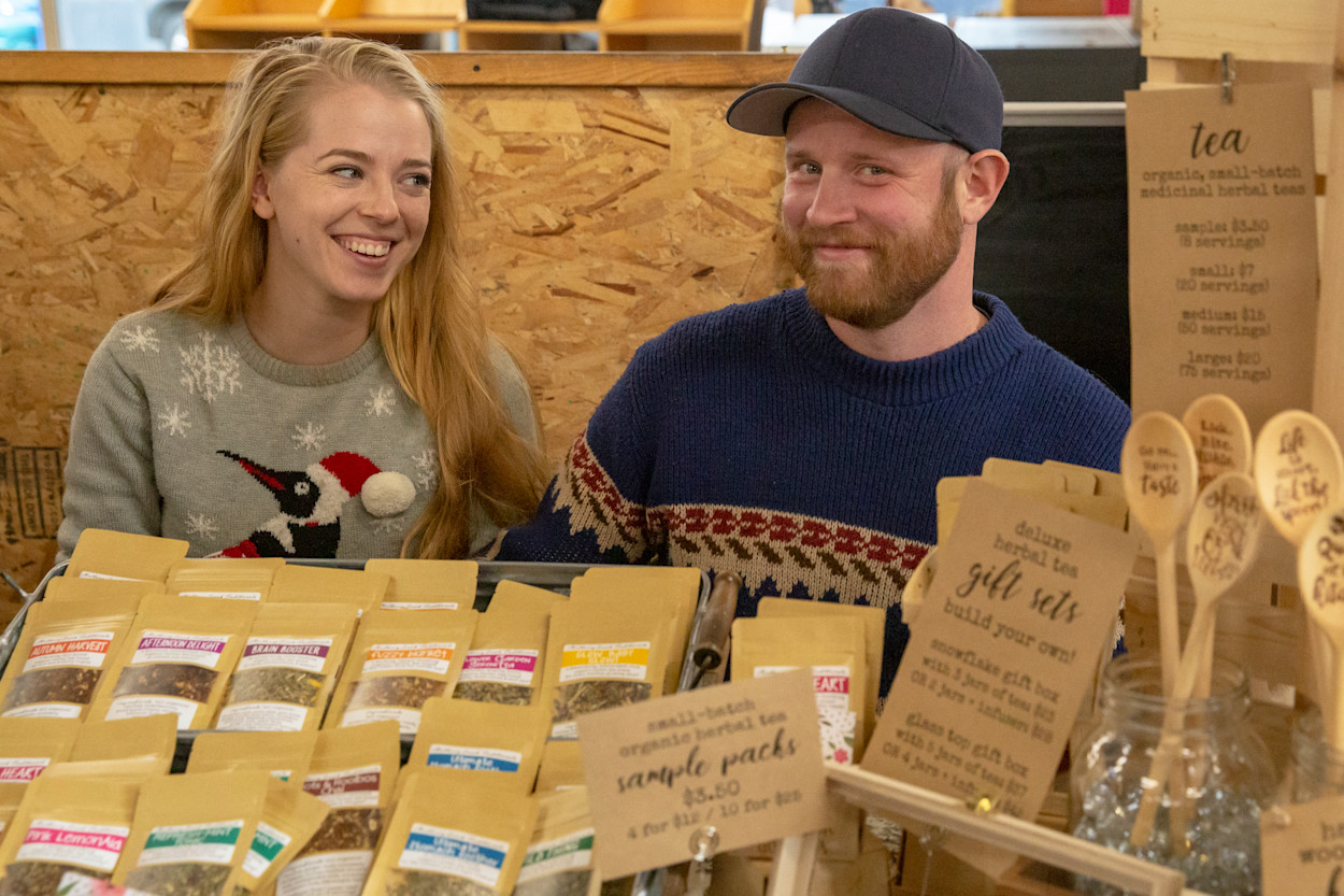 Two grown-ups in holiday sweaters grinning at a craft faire table