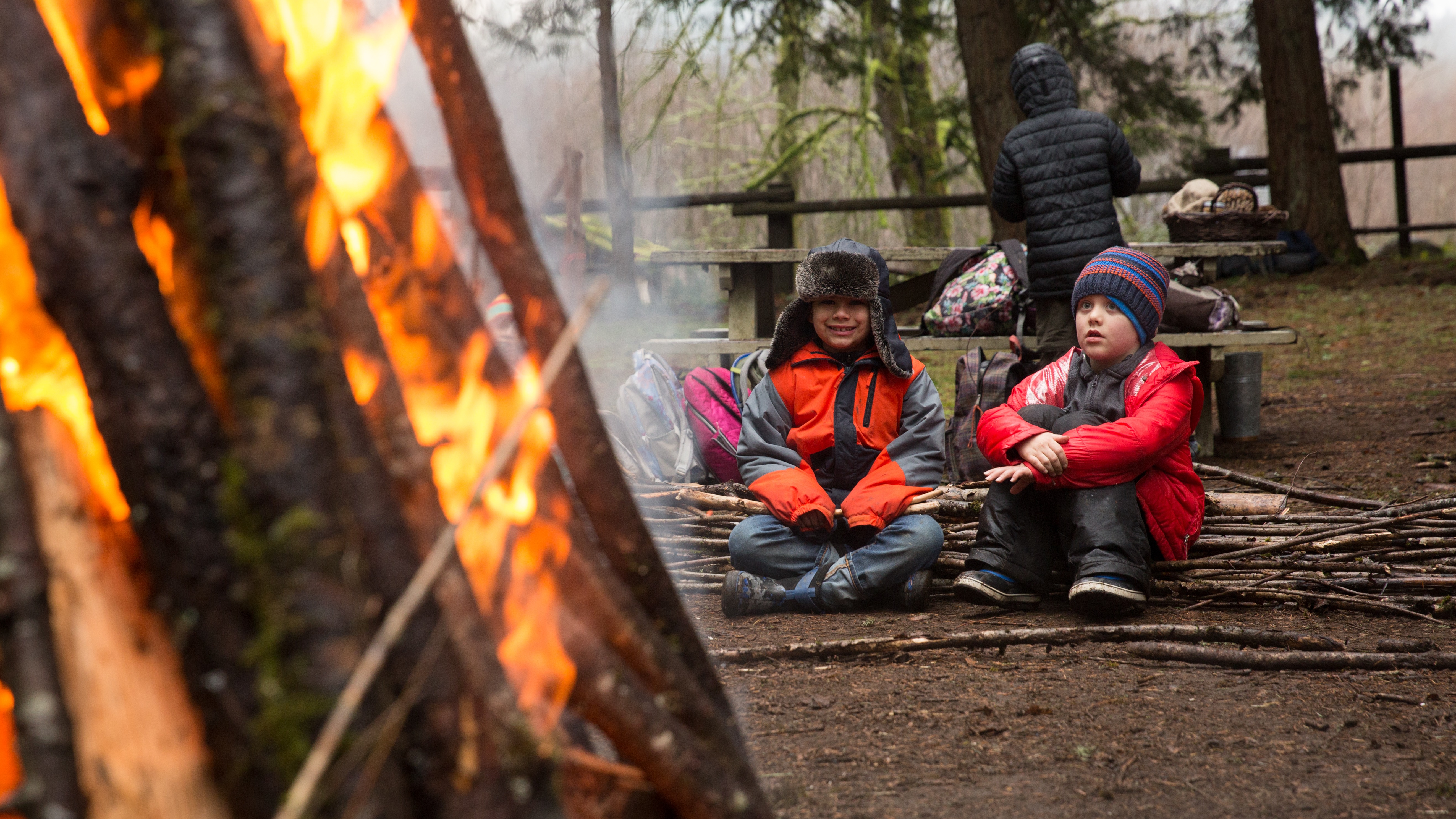 Two bundled-up kids sitting beside a tall winter bonfire