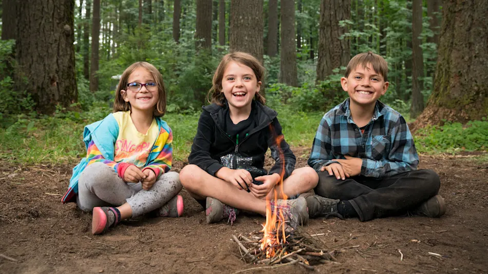 Three kids smiling around a small campfire in the forest