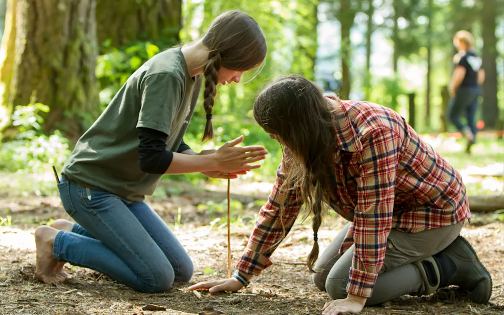 Two A-Team teens working a bow drill in the forest